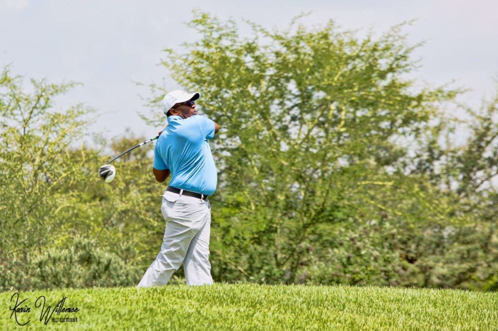 A person in a light blue shirt and white pants swings a golf club on a lush green golf course, surrounded by trees and a blue sky.